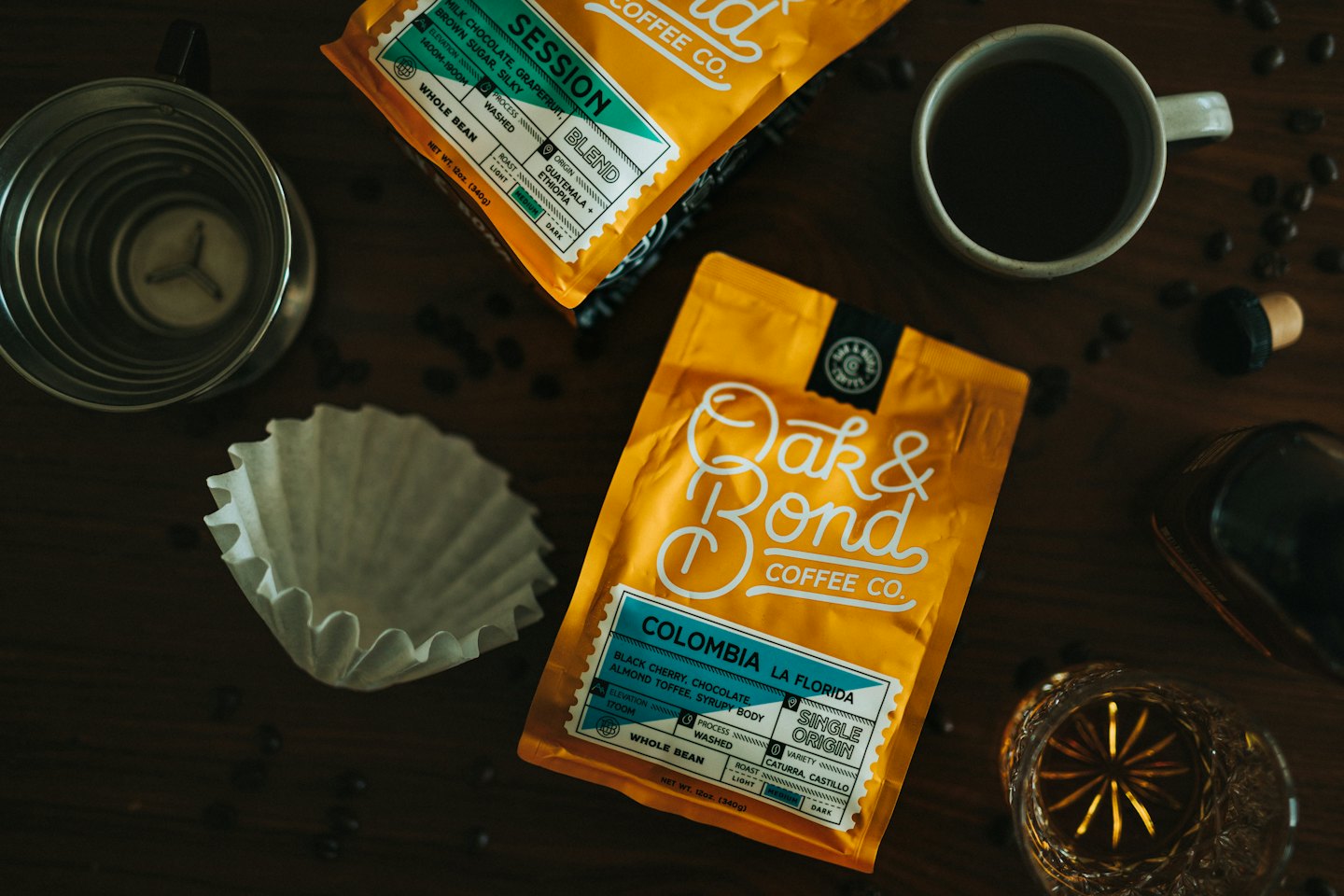 Two bags of coffee beans surrounded by brewing equipment on wood.